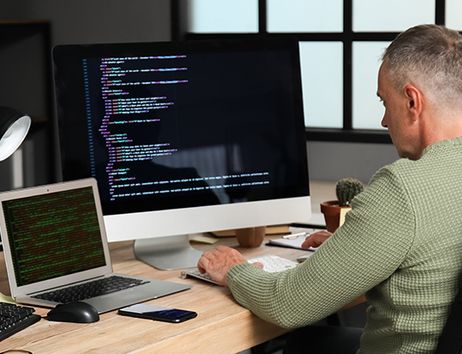 Es ist ein Mitarbeiter vor einem großen Monitor sowie einem Laptop zu sehen