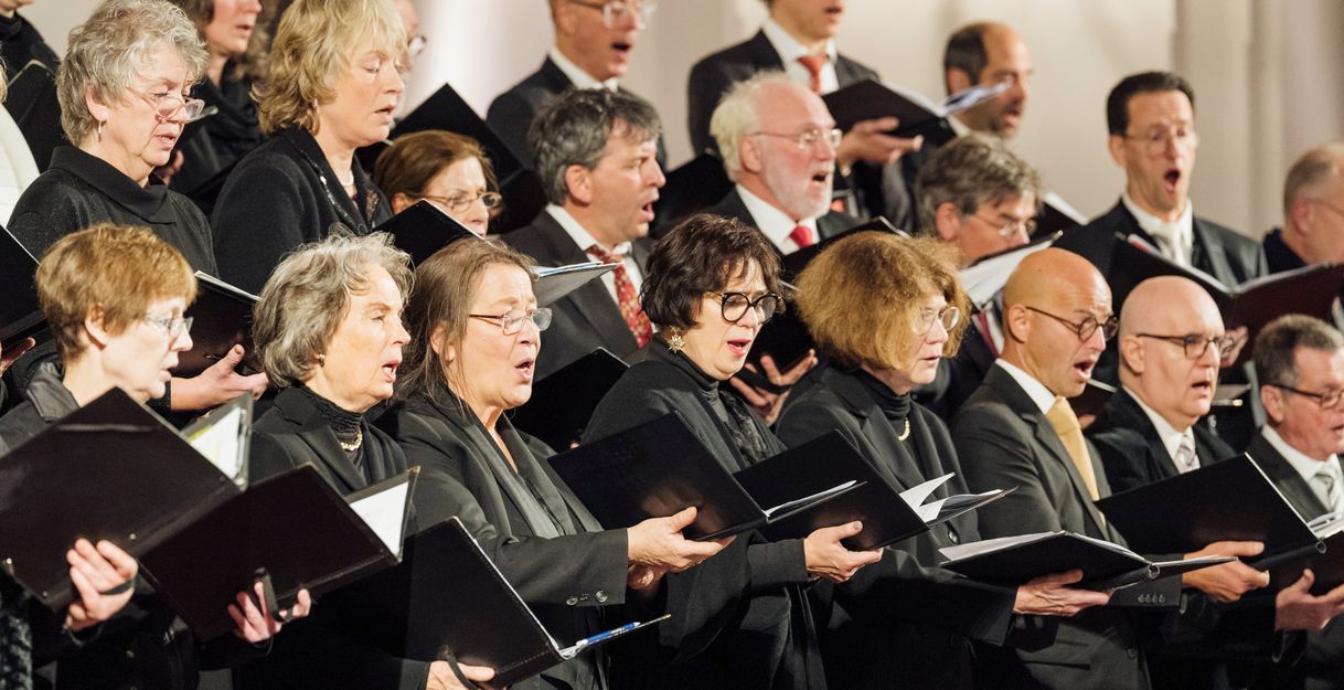 Ein Chor singt in einem Konzert, die Mitglieder tragen schwarze Kleidung und halten Notenblätter.