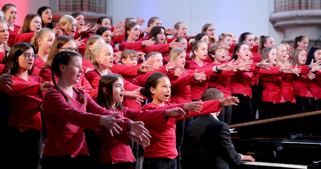 Ein Mädchenchor in roten Blusen singt mit ausgebreiteten Armen auf einer Bühne.