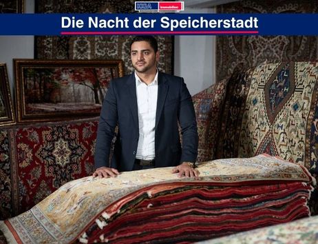 Ein Mann steht in einem Raum voller orientalischer Teppiche, die aufgestapelt sind. Im Hintergrund sind gerahmte Bilder zu sehen.