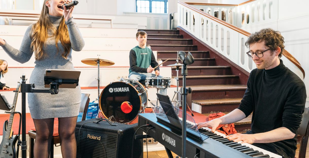 Eine junge Band probt in einer Kirche. Eine Sängerin singt ins Mikrofon, während ein Mann Keyboard spielt und ein anderer Schlagzeug spielt.