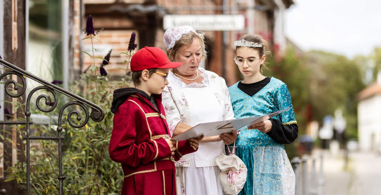 Eine Frau in historischer Kleidung zeigt zwei Kindern in Kostümen eine Karte während einer Stadtführung in Eutin.