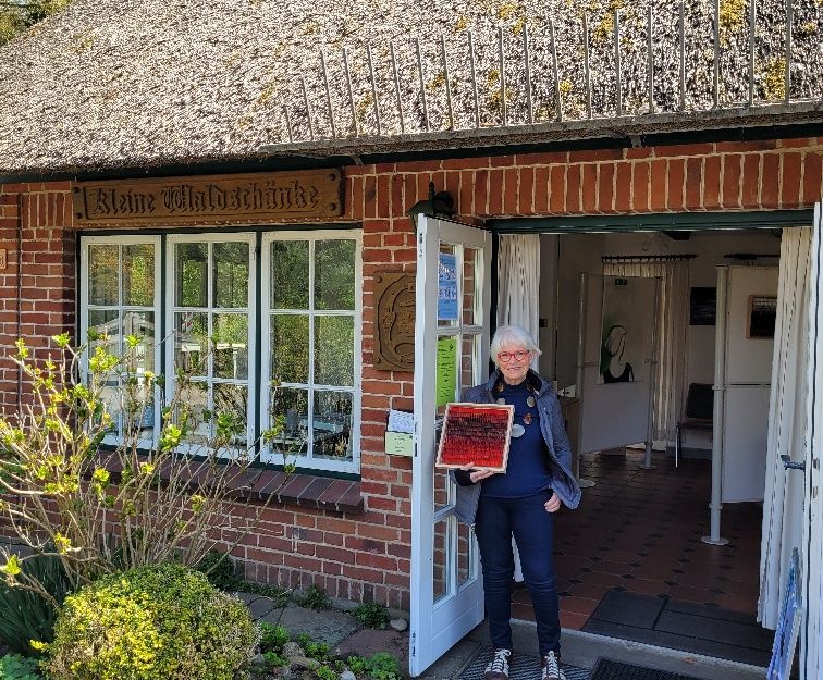 Eine Person steht vor einem Backsteinhaus mit Reetdach und hält ein Kunstwerk in der Hand. Über der Tür steht 'Kleine Waldschänke'.