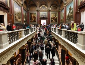 Menschenmenge in einem kunstvoll dekorierten Museum mit Gemälden an den Wänden.