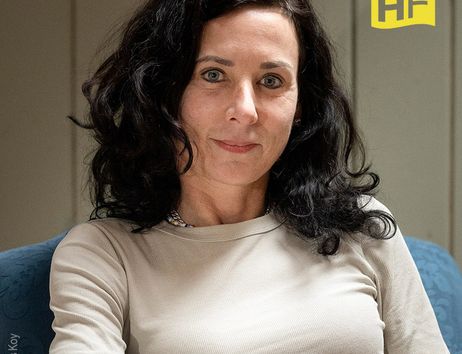 Porträt einer Frau mit dunklen Haaren in einem beigen Oberteil, sitzend auf einem blauen Stuhl. Im Hintergrund ist ein HF-Logo zu sehen.