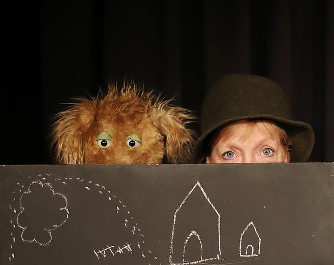 Eine Person mit Hut und eine zottelige Hunde-Handpuppe schauen hinter einer Tafel mit Kreidezeichnungen eines Hauses und eines Baumes hervor.
