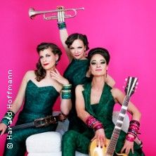 Drei Frauen in grünen Outfits posieren mit Musikinstrumenten vor einem pinken Hintergrund.