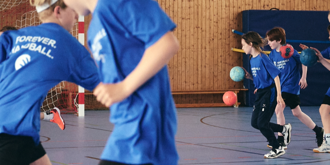 Kinder in blauen T-Shirts, die einen Handball prellend durch eine Sporthalle laufen