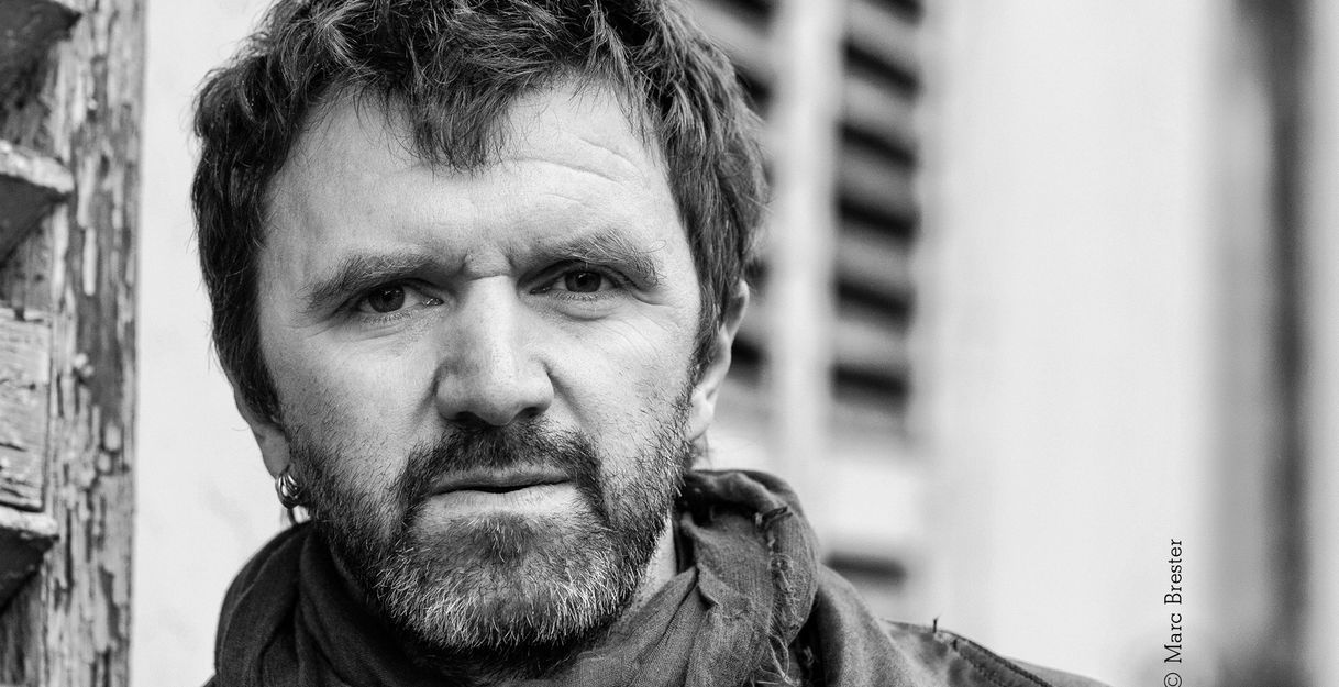 Schwarz-weißes-Portraitfoto einer männlich gelesenen Person mittleren Alters mit Bart. Sie trägt einen Schal, mehrere Ohrringe im einen Ohr und schaut ernst.