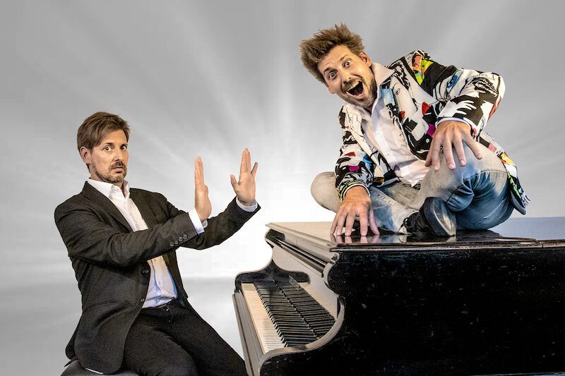 Zwei Männer an einem Flügel, einer sitzt auf dem Klavier, der andere daneben mit erhobenen Händen.