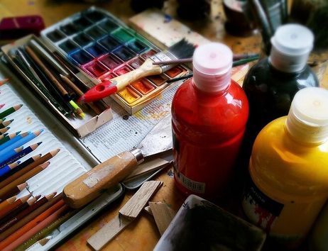 Ein Tisch voller Malutensilien wie Buntstifte, Wasserfarben, Pinsel und Flaschen mit Acrylfarbe in verschiedenen Farben.