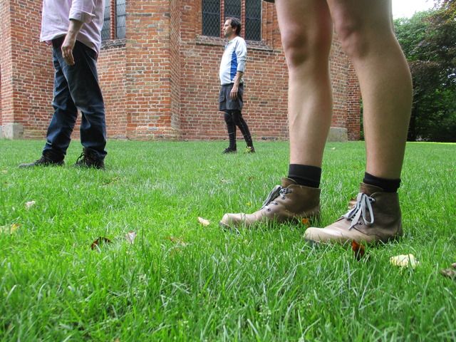 Drei Personen stehen auf einer grünen Wiese vor einem Backsteingebäude.