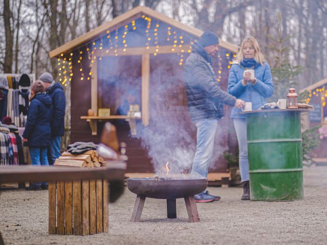 Menschen auf einem Weihnachtsmarkt im Freien mit Feuerstelle und Verkaufsständen.