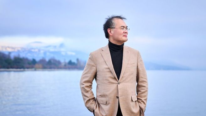 Ein Mann in einem beigen Anzug steht vor einem See mit Bergen im Hintergrund.
