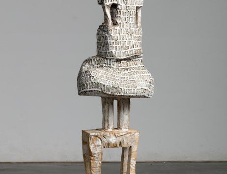 An abstract sculpture made of wood that represents a human figure. It has a round head and a textured surface.