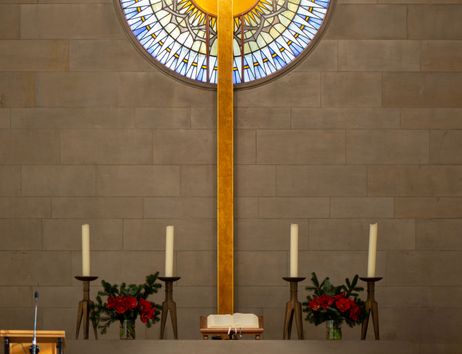 Kirchenaltar mit großem Holzkreuz und buntem Glasfenster im Hintergrund.