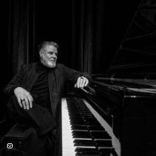 Schwarz-weiß Foto eines Mannes, der lässig an einem Flügel sitzt.