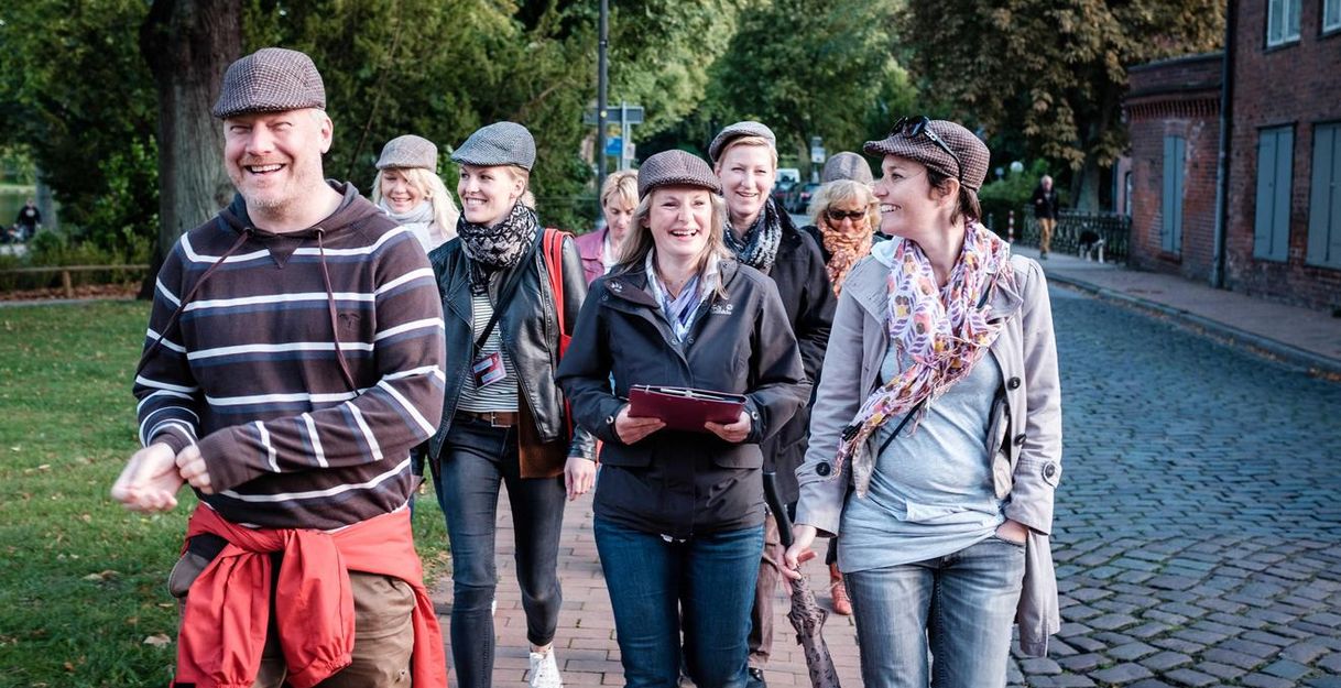 Eine Gruppe von Menschen mit Hüten geht lächelnd auf einem gepflasterten Weg in einer grünen Umgebung.