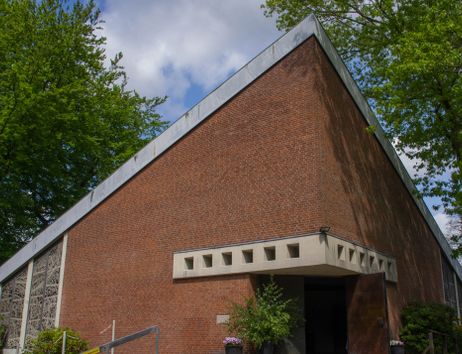 Eine moderne Backsteinkirche mit schrägem Dach und geometrischen Fenstern, umgeben von Bäumen.