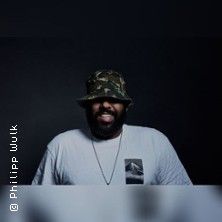 Person mit Hut und Bart vor dunklem Hintergrund.