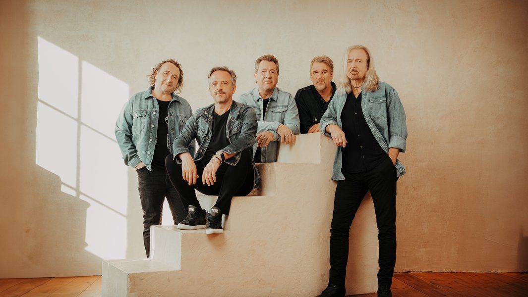 Fünf Männer in Jeansjacken posieren auf einer Treppe in einem hellen Raum.