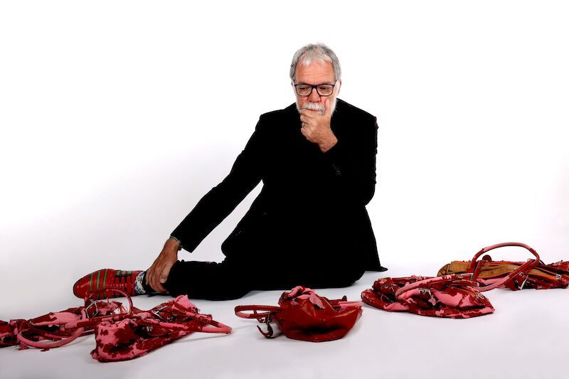 Ein Mann in schwarzer Kleidung sitzt auf dem Boden und betrachtet nachdenklich mehrere rote Handtaschen um ihn herum.