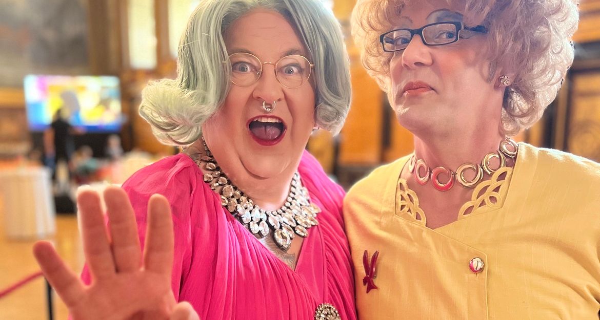 Zwei Dragqueens in bunten Outfits posieren fröhlich in einem eleganten Raum.