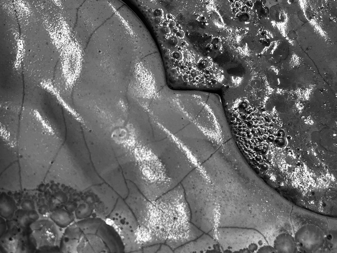 Nahaufnahme in Schwarz-Weiss: glänzende, rissige Oberflächen mit blasenartigen Strukturen und organisch geschwungenen Formen, die an Keramikglasur oder erstarrte Flüssigkeit erinnern.