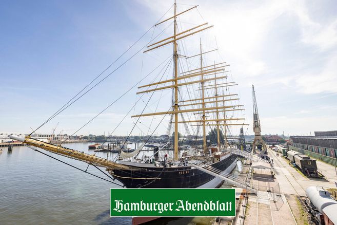 Das Segelschiff Peking liegt im Hafen, umgeben von Kränen und Gebäuden, unter einem klaren blauen Himmel.