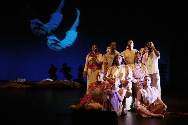 Eine Gruppe von Performerinnen in beigen und roten Kostümen sitzt und steht in einer Formation auf der Bühne. Im Hintergrund sind Silhouetten von Musikerinnen und eine Projektion eines schwebenden Körpers zu sehen.
