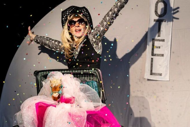 Eine Person in einem glitzernden Outfit und Cowboyhut steht hinter einem Koffer mit einem Spielzeugpony, umgeben von Konfetti.