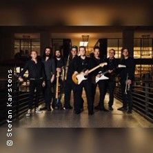 Eine Gruppe von acht Musikern mit Instrumenten in einem beleuchteten Innenraum.