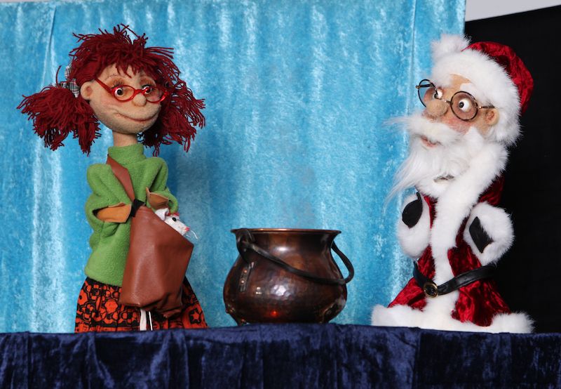Zwei Puppen, eine als Weihnachtsmann und eine mit roten Haaren, stehen vor einem Kupferkessel auf einer Bühne.