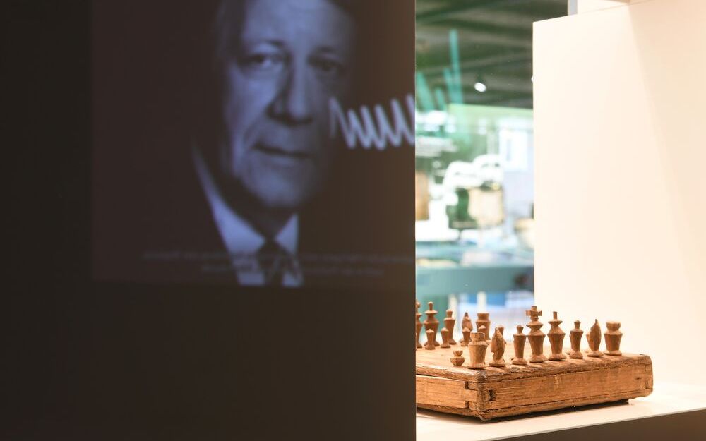 Foto von Wand, auf die ein Foto von Helmut Schmidt projeziert ist. Die Wand enthält einen Durchbruch, in dem im hellen Licht ein abgenutztes Schachspiel aus hellem Holz steht.