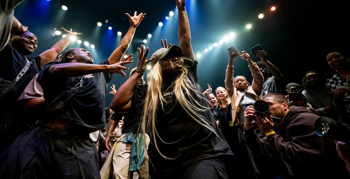 A dynamic scene in which a crowd of people stand around a dancer, cheering and cheering her on with gestures. The dancer raises her arms upwards, spreading her fingers. At the bottom right, someone photographs the dancer from close up.