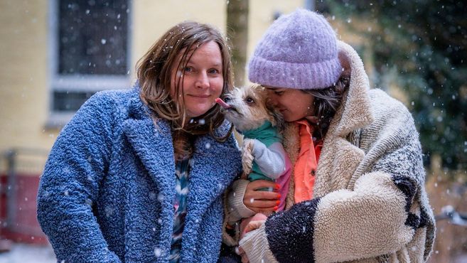 Zwei Frauen in Winterkleidung halten einen kleinen Hund im Schnee.