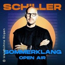 Ein Mann mit Brille steht vor einem orange-blauen Hintergrund mit den Worten 'SCHILLER SOMMERKLANG OPEN AIR'.