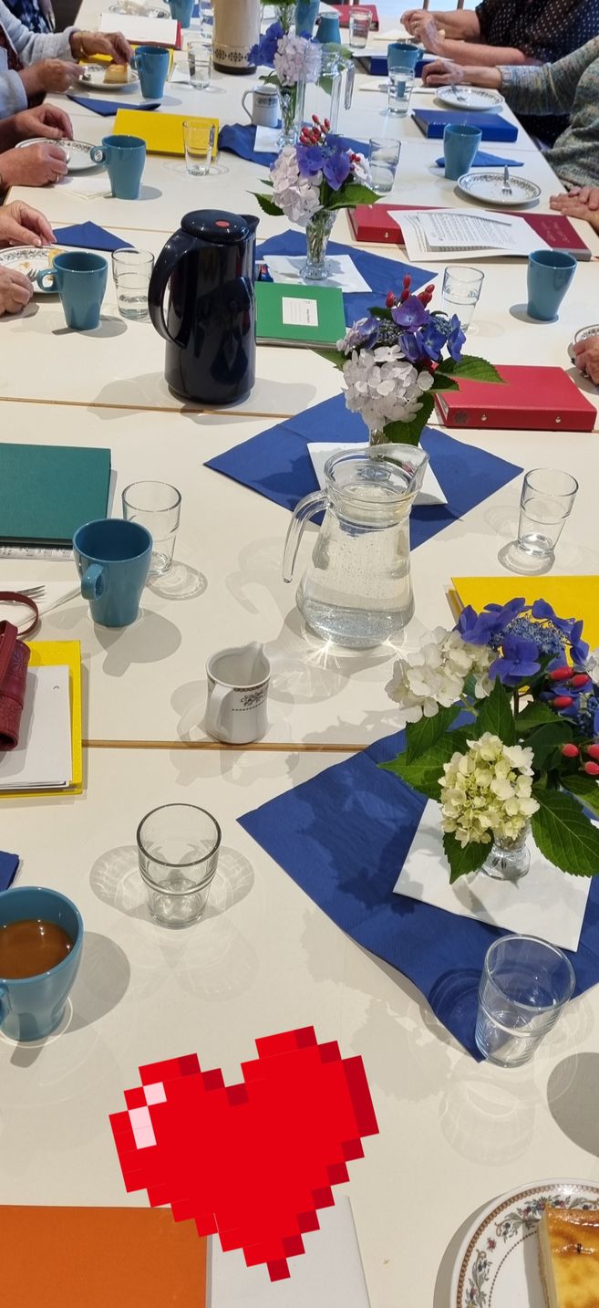 Tisch im Seniorenclub mit älteren Personen, blauen Tassen, Wasserkrügen, Blumenvasen und Kuchen bei einer gemeinsamen Kaffeerunde.