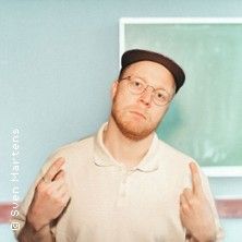 Ein Mann mit Brille und Mütze steht vor einer Tafel und zeigt mit beiden Händen nach oben.