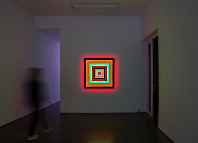 Ausstellungssaal mit einer leuchtenden Neon-Installation aus konzentrischen Quadraten an der Wand; links geht eine unscharfe Person vorbei.