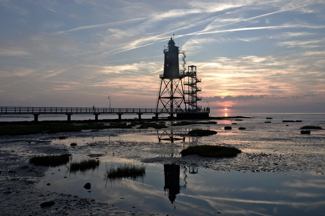 Leuchtturmdenkmal Obereversand bei Sonnenuntergang mit Steg, Spiegelungen im Watt und weitem Himmel.