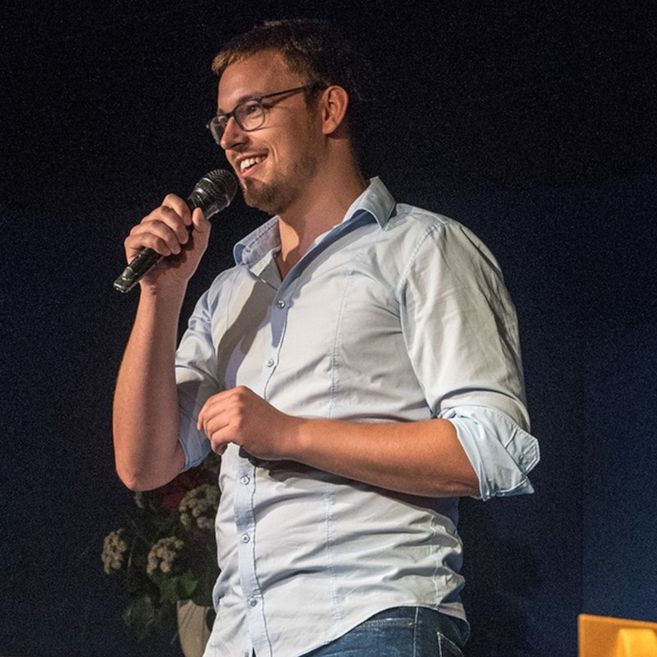 Ein Mann mit Brille und Bart hält ein Mikrofon und lächelt auf einer Bühne.