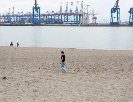 Eine Person geht barfuß am Sandstrand entlang, im Hintergrund sind Hafenkräne zu sehen.