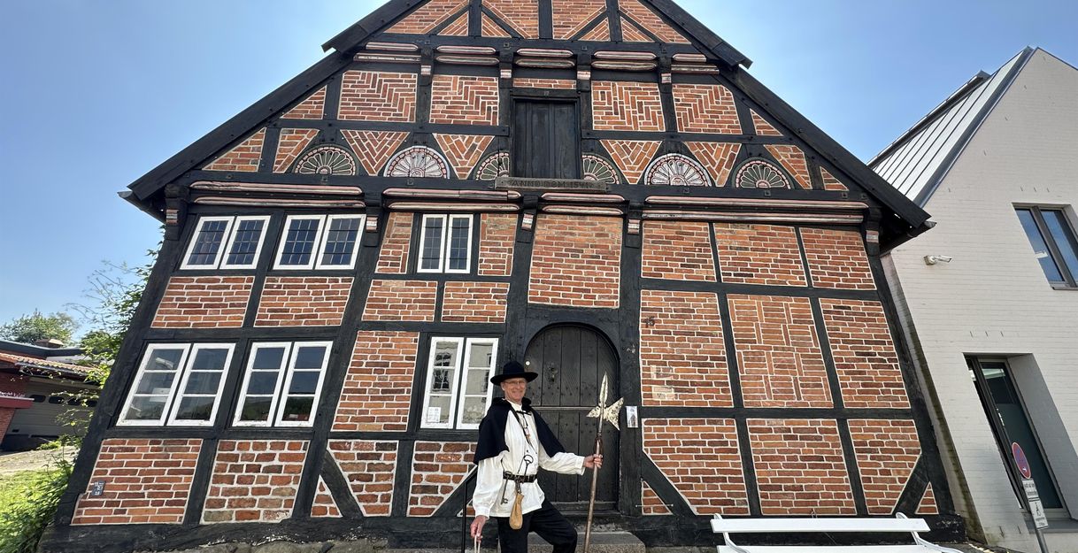 A historic half-timbered house with red bricks and black timber details. A person in traditional clothing stands in front of it.