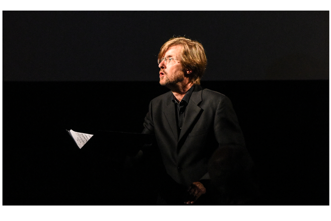 Ein Mann in einem dunklen Anzug hält ein Notenblatt und steht vor einem dunklen Hintergrund.