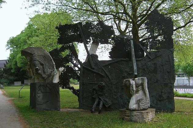 Foto eines Denkmals für den 2. Weltkrieg, das aus drei Teilen besteht. Zwei kleinere abstrakte Elemente aus Stein stehen je auf einem Steinsockel. Das größte Element ist schwarz, flächig und eine Person ragt mit Beinen und Mantel heraus, ebenso Stangen.