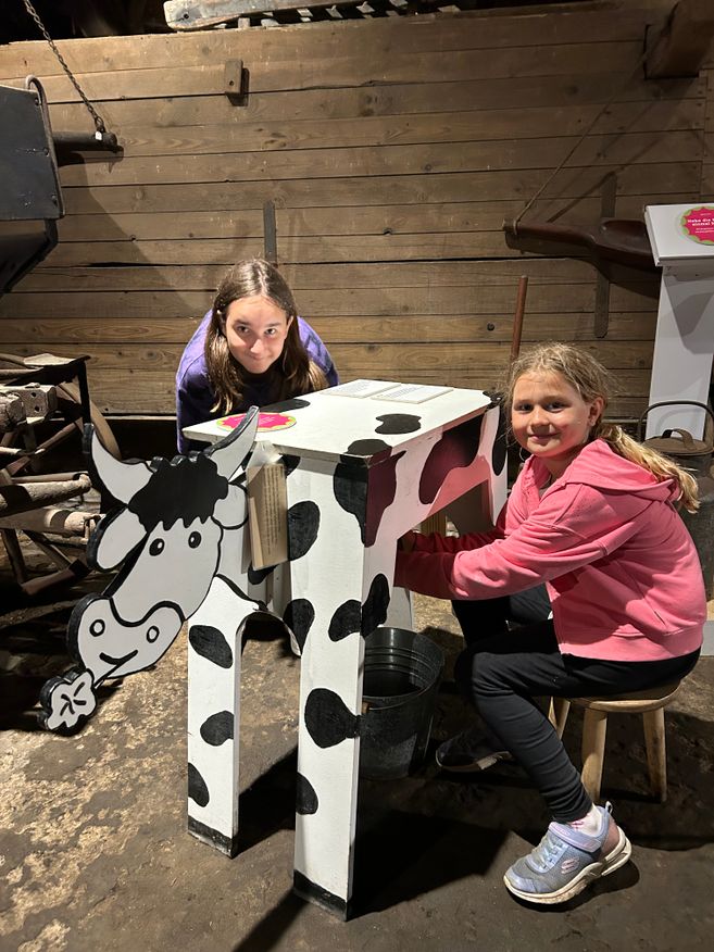 Zwei Ferien-Kinder sitzen auf dem Melkschemel und melken die Holzkuh 