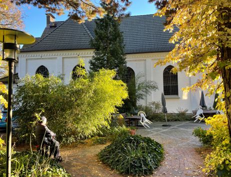Ein herbstlicher Garten mit einer weißen Kirche im Hintergrund. Eine Person sitzt in einem Rollstuhl und genießt die Sonne.
