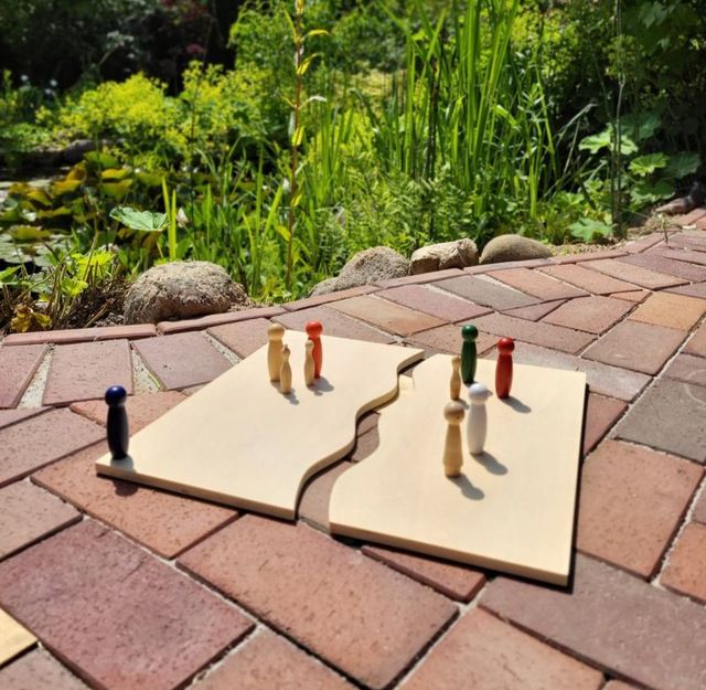 Holzfiguren auf zwei Holzplatten im Freien, umgeben von Pflanzen.