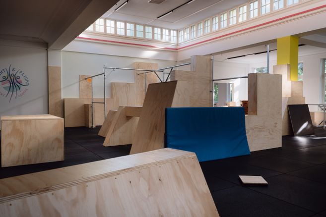 In der hellen Parkourhalle mit Oberlichtern und dunklem Gummi Boden finden sich unterschiedlich Holzhindernisse und Stangen für das Parkourtraining.